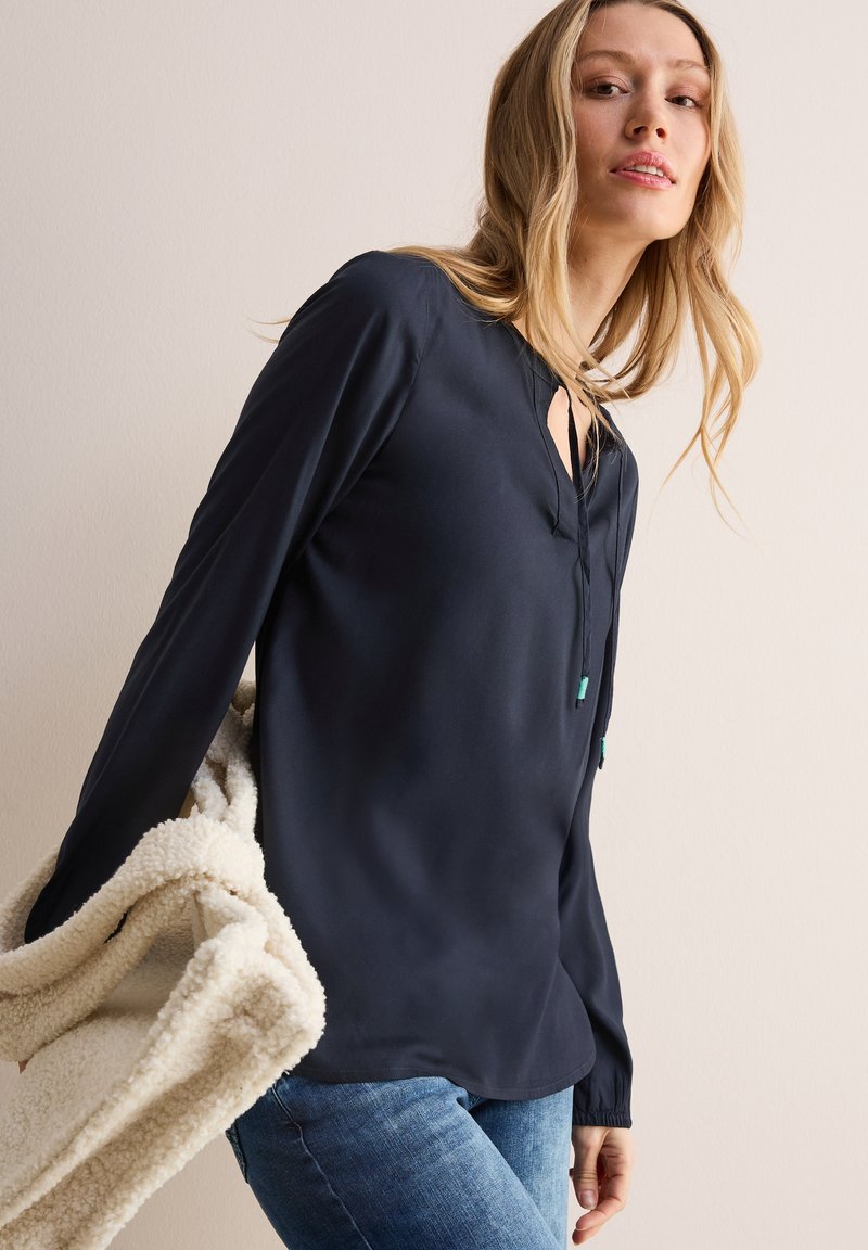 Marineblauwe blouse met lange mouwen en een keyhole-halslijn, soepele textuur en een ontspannen pasvorm, gecombineerd met een crèmekleurige, harige sjaal.