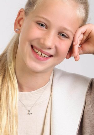 Zilveren hartvormige oorbellen en een bijpassende halsketting bij een kind met lichtblond haar. Draagt een beige trui onder een lichtbruine cardigan.