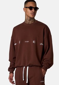 Brown sweatshirt with white graphic text on the front, featuring long sleeves and a relaxed fit, paired with coordinated trousers.