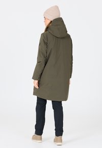 Olivgrön lång kappa med huva, har en mjuk textur och framfickor. Tillsammans med mörka byxor och beige ankelstövlar.