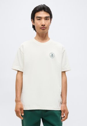 Νέος άνδρας με μαύρα μαλλιά που φοράει λευκό μπλουζάκι με μικρό κυκλικό λογότυπο στο αριστερό στήθος και πράσινο παντελόνι.