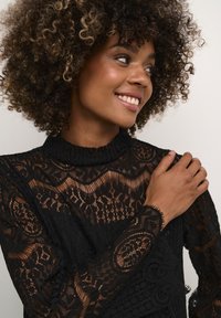 Black lace top featuring intricate patterns, ruffled neckline, and textured sleeves. Curly hair frames the model's smiling face.