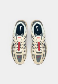 Par bež i sivih Nike tenisica s crvenim naglascima postavljenih jedan pored drugog, gledano odozgo na bijeloj podlozi.