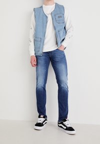 Jeansväst med fickor, lager på lager över en vit sweatshirt, tillsammans med blå jeans och svarta sneakers med vita detaljer.