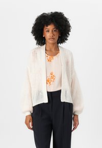Cardigan deschis la culoare, din tricot ușor, cu o croială lejeră și mâneci tip balon, combinat cu o bluză cu imprimeu floral și pantaloni negri.