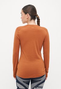 Burnt orange long-sleeve shirt with a smooth texture, rounded neckline, and fitted design. Paired with patterned dark leggings featuring geometric designs.