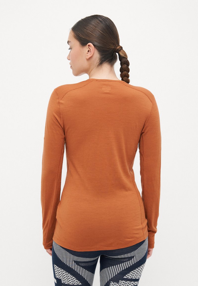 Gebranntes orangefarbenes Langarmhemd mit glatter Textur, rundem Ausschnitt und taillierter Passform. Kombiniert mit gemusterten dunklen Leggings mit geometrischen Designs.