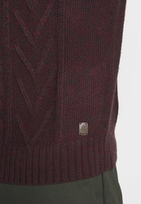 Bordeauxfarbener Strickpullover mit einem Kabelmuster, geripptem Saum und einem kleinen metallischen Logoschild an der Seite. Strukturiertest Gewebe mit subtilen Farbvariationen.
