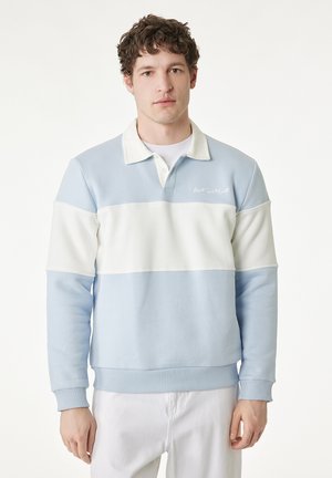 Młody mężczyzna o kręconych, brązowych włosach, ubrany w jasno niebiesko-białą, kolorową, długorękawową koszulkę polo i białe spodnie, stojący zwrócony przodem.