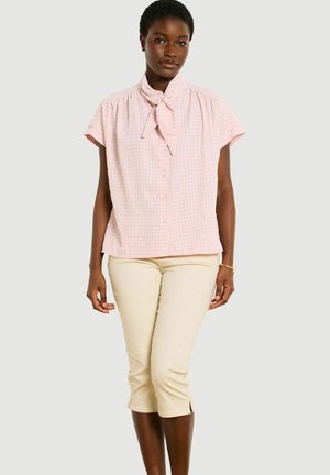 Kobieta w różowej bluzce z krótkim rękawem i krawatem oraz beżowych krótkich spodniach, stojąca na jednolitym jasnoszarym tle.