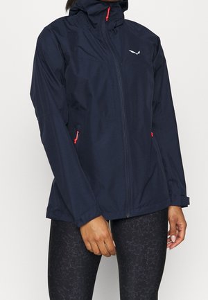 Personne portant une veste zippée bleu marine avec des tirettes rouges et des leggings noirs à motifs, debout devant un fond blanc uni.