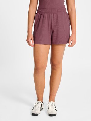 Kvinde model iført mauve elastiske shorts med høj talje og hvide sneakers med sorte detaljer, stående mod en ensartet lys baggrund.