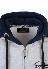 Grijze en marineblauwe zip-up hoodie met een gevoerde capuchon, trekkoorden en een ritssluiting aan de voorkant. Bevat een logo en tekst aan de binnenkant van de kraag.
