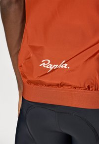 Jachetă de ciclism roșie cu o textură netedă, având un logo alb "Rapha." la margine. Şorturi închise la culoare, ajustate, cu detalii de cusătură vizibile.