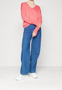 Korálový svetr s výstřihem do V a spadlými rameny, zkombinovaný s širokými modrými jeansy. Model má na sobě bílé tenisky; hladké textury a ležérní střih.