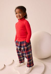 Pulover roșu cu guler înalt, asortat cu pantaloni în carouri navy și roșu, având o croială lejeră și adidași albi. Textură netedă și culori vibrante.