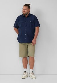 Marine-blauwe overhemden met korte mouwen en twee borstzakken, khaki shorts en witte sneakers met grijze accenten. Lichte achtergrond.