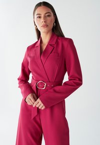 Rožnato-modri kombinezon s pasom, dolgimi rokavi, notch ovratnikom in enim žepom na prsih. Izdelan iz gladkega, strukturiranega blaga.