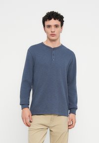 Μπλούζα με μακριά μανίκια henley σε σκούρο μπλε, με στρογγυλή λαιμόκοψη, πατιλέτα με τρία κουμπιά και ριμπ μανσέτες. Φοράται με μπεζ παντελόνια.