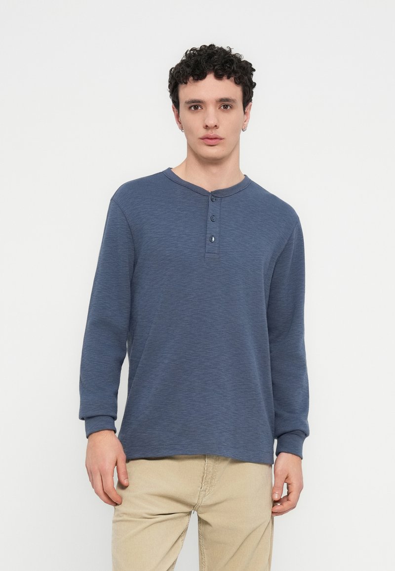 Μπλούζα με μακριά μανίκια henley σε σκούρο μπλε, με στρογγυλή λαιμόκοψη, πατιλέτα με τρία κουμπιά και ριμπ μανσέτες. Φοράται με μπεζ παντελόνια.