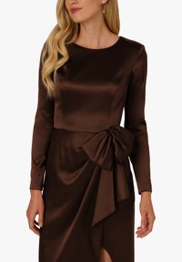 Brązowa satynowa suknia wieczorowa z długimi rękawami, dopasowaną górą i akcentem w postaci bocznej kokardy. Posiada drapowany przód i rozporek sięgający do ud.