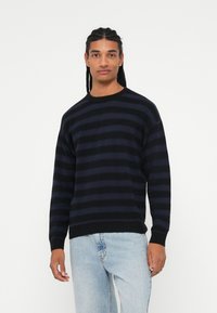 Pull en tricot rayé marine avec un col rond, présentant des lignes horizontales texturées, des manches longues et un ourlet noir, associé à un jean clair.