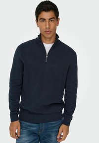 Un sweat zippé bleu marine avec un motif herringbone texturé, doté de poignets et d'un ourlet côtelés. Associé à un jean en denim.
