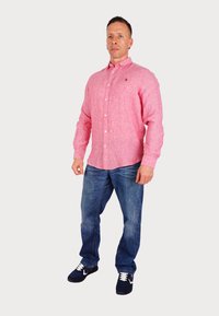 Pink button-up shirt with textured fabric, paired with blue jeans and black sneakers. Shirt features a small logos, creating a casual look.