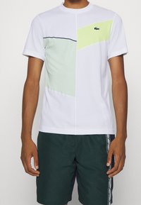Vit kortärmad sporttröja med geometriska paneler i mintgrön och neongul, rund hals, med kontrasterande svart söm och logotyp.
