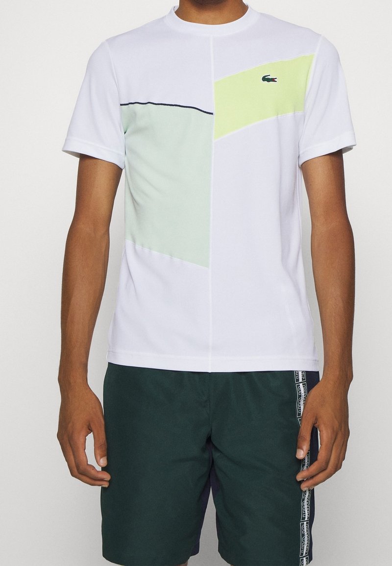 Vit kortärmad sporttröja med geometriska paneler i mintgrön och neongul, rund hals, med kontrasterande svart söm och logotyp.