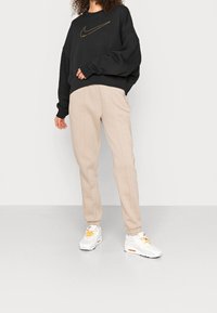 Black sweatshirt with gold Nike logo, beige sweatpants with elastic waistband, and white sneakers with orange accents. Soft fabric texture.