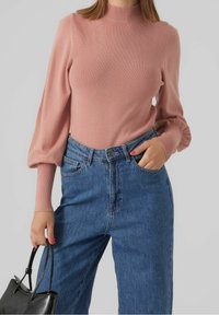 Maglione rosa a coste con alto collo alla coreana e maniche a sbuffo, abbinato a jeans in denim blu a vita alta. Borsa nera in mano.