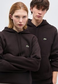 Δύο νέοι ενήλικες που φορούν μαύρα φούτερ Lacoste, στέκονται κοντά ο ένας στον άλλο μπροστά σε ένα απλό ανοιχτόχρωμο φόντο, κοιτάζοντας ευθεία μπροστά.