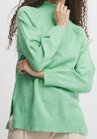 Pull vert menthe avec un col montant, des poignets et un ourlet côtelés. Comprend des fentes latérales pour faciliter les mouvements. Tissu doux et texturé.