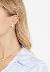 Des boucles d'oreilles en forme de fleurs dorées et blanches présentent une pierre précieuse claire au centre. Portées avec une chemise rayée bleue et blanche et un collier.