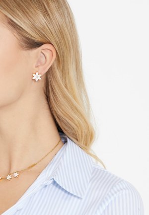 Orecchini a bottone a forma di fiore in oro e bianco con uno strass trasparente centrale. Indossati con una camicia a righe blu e bianche e una collana.