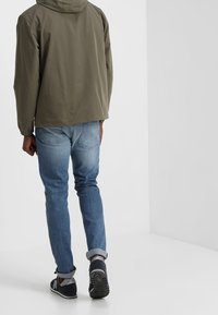 Person som bär en grön huva-jacka, blå uppvikta jeans och svarta sneakers, som går bortåt mot en enkel vit bakgrund.