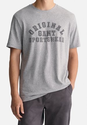 Homme portant un t-shirt gris à manches courtes avec "Original Gant Sportswear" imprimé en noir sur la poitrine et un pantalon foncé.