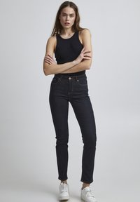 Top negro sin mangas combinado con jeans de mezclilla oscuros de tiro alto. Los jeans tienen cinco bolsillos, herrajes color cobre y detalles de costura en rojo.