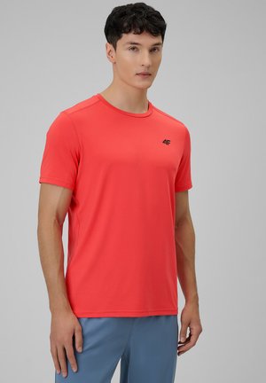 Młody mężczyzna z krótkimi ciemnymi włosami, ubrany w zwykły czerwony sportowy t-shirt z krótkim rękawem i jasnoniebieskie spodnie, stoi na szarym tle.