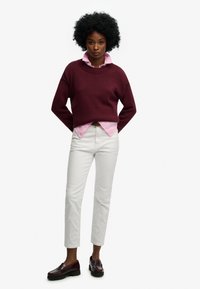 Bordowy sweter z dzianiny na jasnoróżowej koszuli z kołnierzykiem, zestawiony z białymi jeansami o prostych nogawkach i ciemnogranatowymi mokasynami.