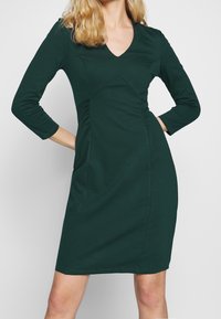 Green, fitted dress with three-quarter sleeves, V-neck, and side ruching. Smooth fabric with a solid color and knee-length hem.