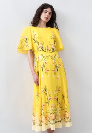 Γυναίκα που φοράει κίτρινο φόρεμα μέσης με φλοράλ σχέδιο και φαρδιά μανίκια με βολάν, στέκεται μπροστά σε απλό ανοιχτό γκρι φόντο.