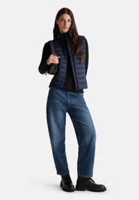 Žena v námořnicky modré prošívané vestě, černém dlouhém rukávu, modrých džínách a černých botách, držící černou kabelku na rameni, stojí na bílém pozadí.