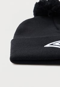 Melnā adīta cepure ar salocītu malu un pomponu augšpusē, uz malas ir balta izšūta ģeometriska atzīme.