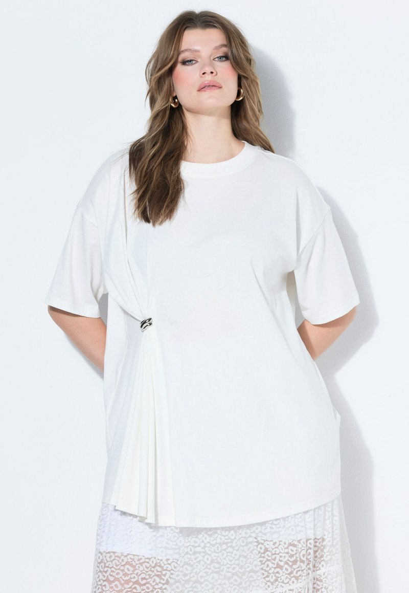 Frau mit welligem braunem Haar, die große Creolen trägt, ein locker sitzendes weißes Oberteil mit seitlichen Falten, befestigt durch einen Metallring, und einen weißen Spitzenrock.