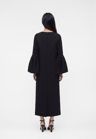 Γυναίκα με μακριά σκουρόχρωμα μαλλιά, που φοράει μαύρο midi φόρεμα με μακριά μανίκια που φουσκώνουν και μαύρα ψηλοτάκουνα παπούτσια με λουράκι στον αστράγαλο, απεικονιζόμενη από πίσω.