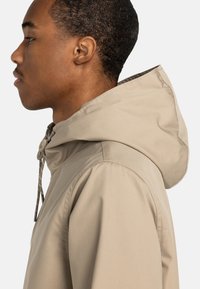 Beige Kapuzenjacke mit Kordelzug und Knopfdetail, die eine glatte Textur und ein einfaches Design aufweist. Von einer Person getragen, seitliche Ansicht.