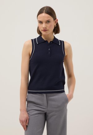 Femme portant un polo sans manches bleu marine avec bordure blanche et un pantalon à fines rayures grises, debout avec une main dans la poche, sur un fond neutre.