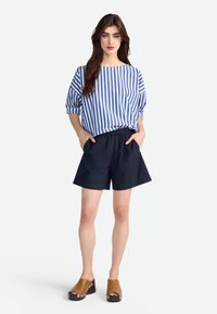 Blouse rayée bleue et blanche avec des manches bouffantes, associée à un short marine et des sabots marron avec des accents en argent décoratifs.